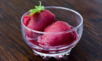 Two scoops of vibrant raspberry sorbet garnished with a mint leaf in a glass bowl.