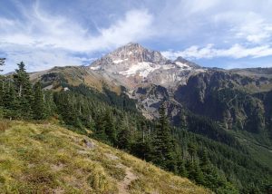 Snow-capped mountain towering over green forested hills under a partly cloudy sky.
