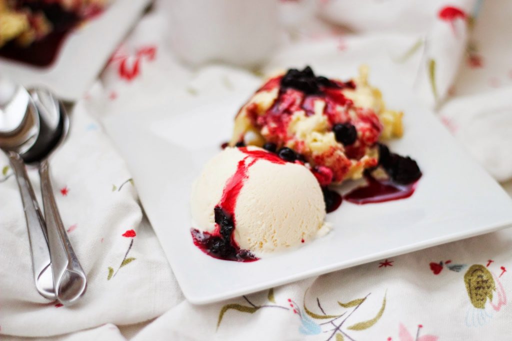 Vanilla ice cream topped with berry sauce and fruit crumble on a white plate.