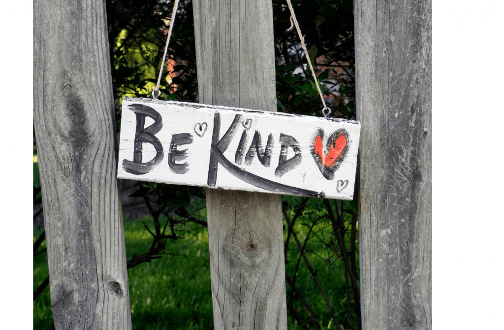 A wooden sign with 'Be Kind' and a red heart hanging on a fence.